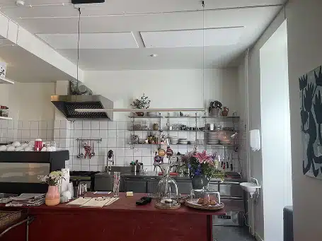 Indblik i et åbent cafékøkken med røde bardiske, flisevægge med hylder fyldt med service, en emhætte over komfuret og friske blomster på disken.