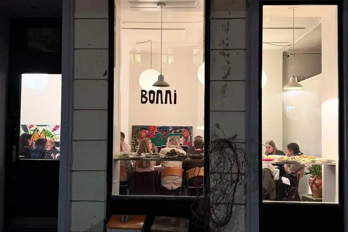 Bonnis facade om aftenen set udefra, hvor gæster sidder til bords og spiser indendørs, oplyst bag store butiksruder med Bonni-logoet.